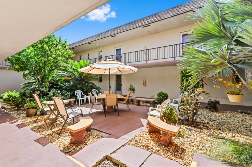 20260916183219887639000000-o - Lake Worth Homes - Lake Worth Homes Outdoor hotel courtyard with a central table under a beige umbrella, surrounded by chairs and potted plants on a gravel garden.
