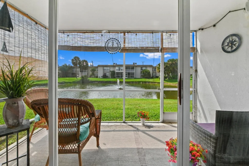 20260916183725202797000000-o 1 - Lake Worth Homes - Lake Worth Homes Screened patio with wicker chairs and a potted plant, overlooking a pond and apartment buildings in a sunny residential view.