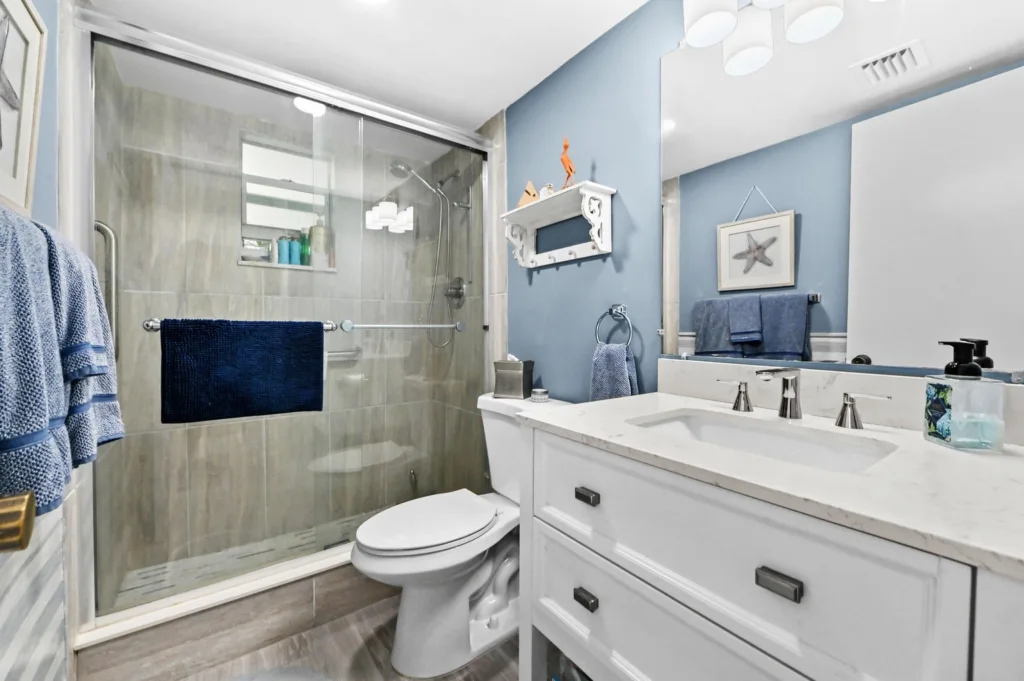 20260916183601388691000000-o - Lake Worth Homes - Lake Worth Homes Blue bathroom with a glass-enclosed shower, white vanity, and navy towels on racks.