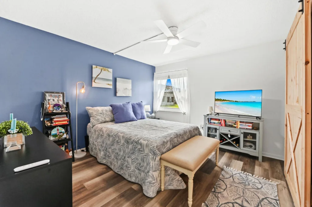 20260916183537511041000000-o - Lake Worth Homes - Lake Worth Homes Bedroom with a blue feature wall, queen bed with a palm-leaf quilt and purple pillows, and a bench at the foot of the bed.