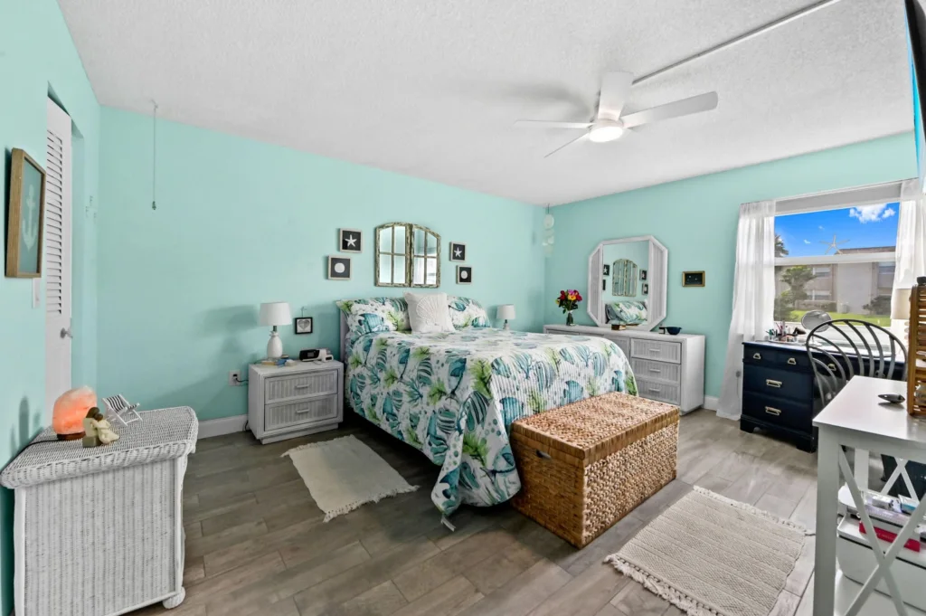 20260916183615033371000000-o - Lake Worth Homes - Lake Worth Homes Coastal-themed bedroom with mint walls, queen bed in tropical quilt, two nightstands, and a wicker storage bench at the foot of the bed.