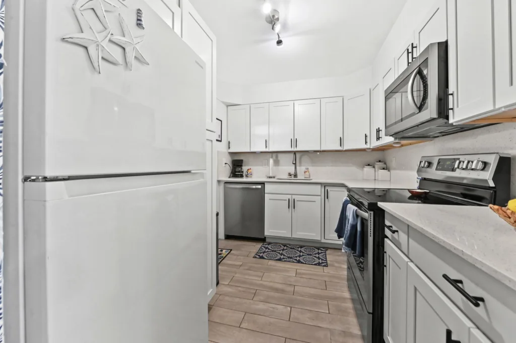 20260916183404890298000000-o - Lake Worth Homes - Lake Worth Homes Modern white kitchen with stainless steel fridge on the left, matching appliances, and light wood-look tile flooring.