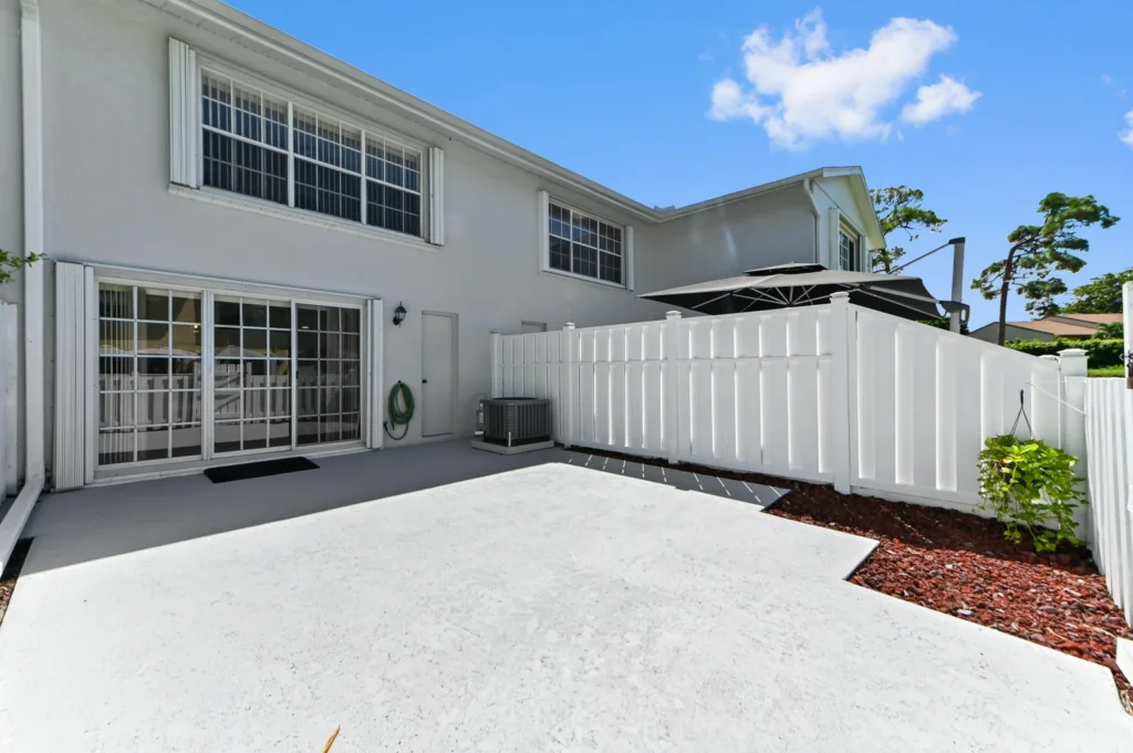 Backyard patio beside a white house with sliding glass doors and a tall white privacy fence under a blue sky with a large umbrella area.