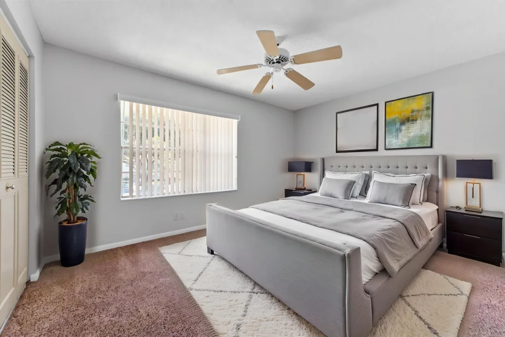 Light gray bedroom with a queen bed, tufted headboard, two dark wood nightstands, and abstract wall art above the bed. A ceiling fan hangs from the ceiling and a potted plant sits near a large window with vertical blinds.