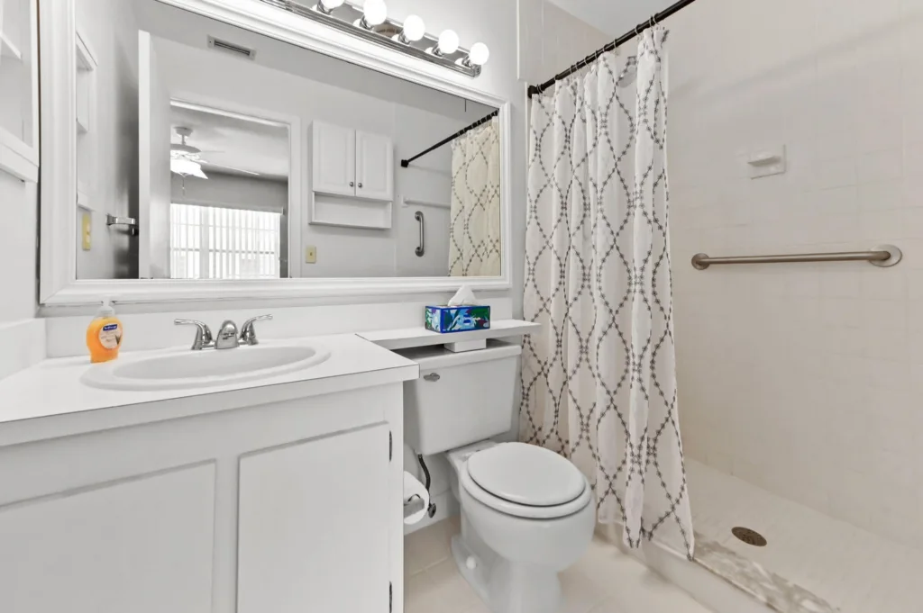 Bright white bathroom with vanity, large mirror, toilet, and a shower with a patterned curtain. A bottle of soap sits on the sink.