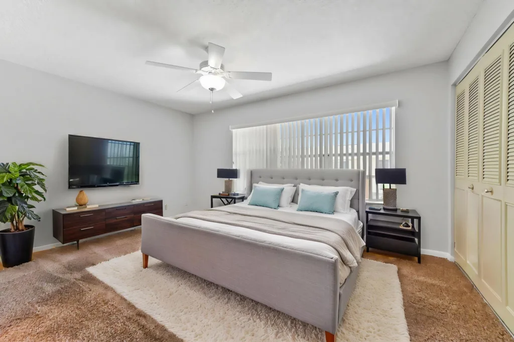 Bright modern bedroom with a gray upholstered king/queen bed, two black nightstands, and a wall-mounted TV on a dark wood dresser under a ceiling fan.