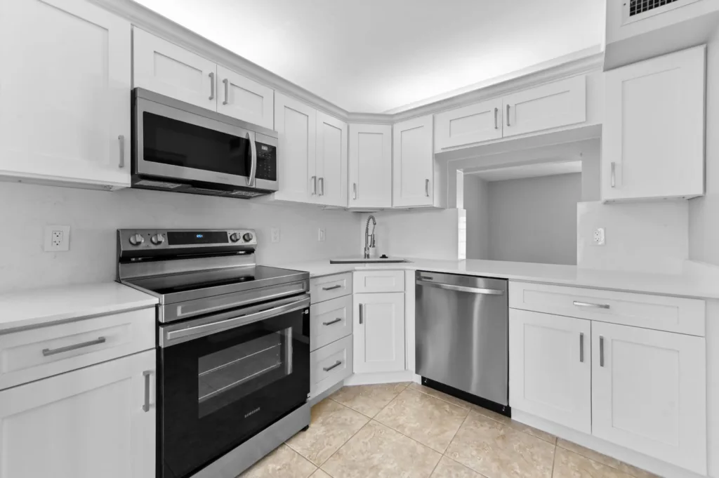 Modern L-shaped kitchen with white cabinets and stainless steel appliances (oven, microwave, dishwasher) and tiled floor—bright and clean.