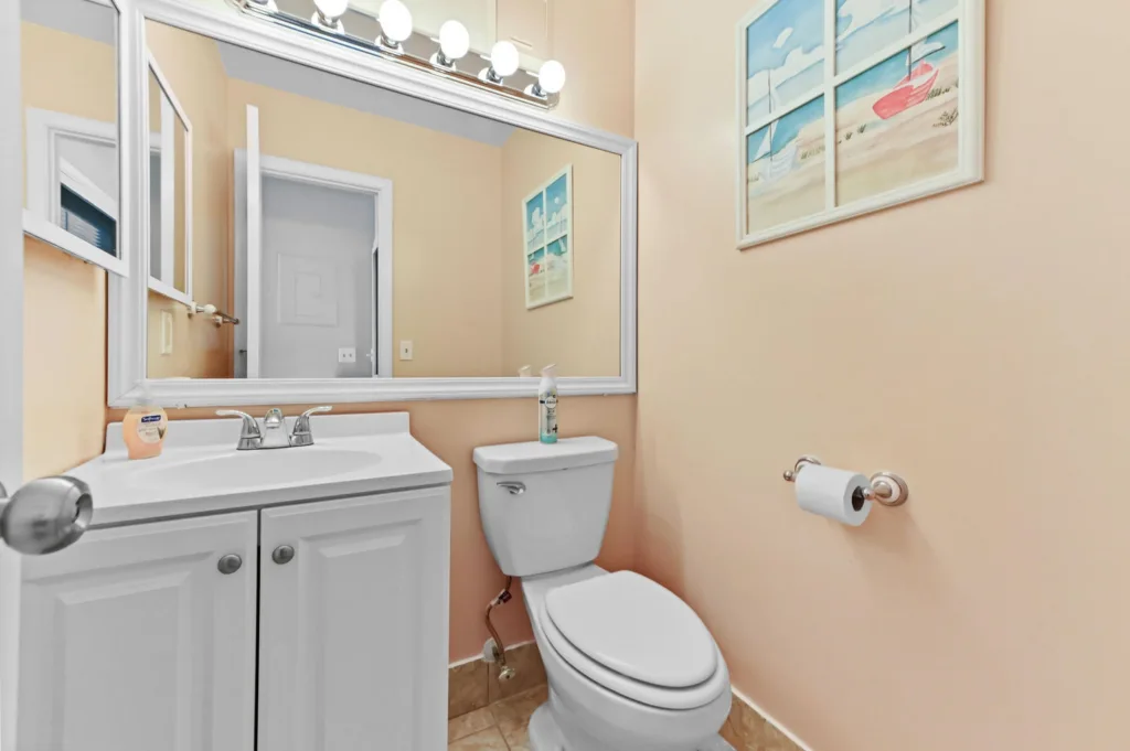 White bathroom vanity with sink and silver faucet, large framed mirror with bright bulbs, and a toilet against beige walls with a toilet paper holder nearby; beach-themed art on the wall.