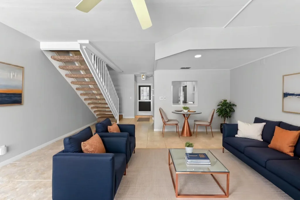 Modern living room with navy sofas, orange throw pillows, and a glass coffee table on a light rug; staircase and dining nook in the background.
