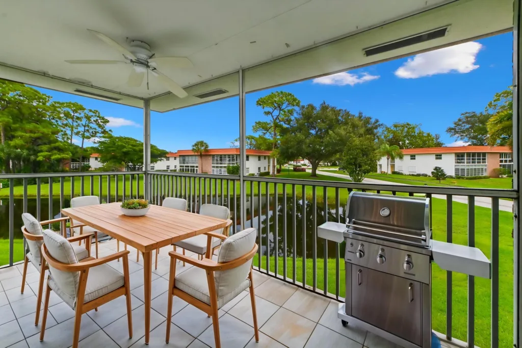 20260923153328352233000000-o - Lake Worth Homes - Lake Worth Homes screened balcony with a wooden dining table, cushioned chairs, and a stainless grill overlooking a green lawn and blue sky nearby apartments