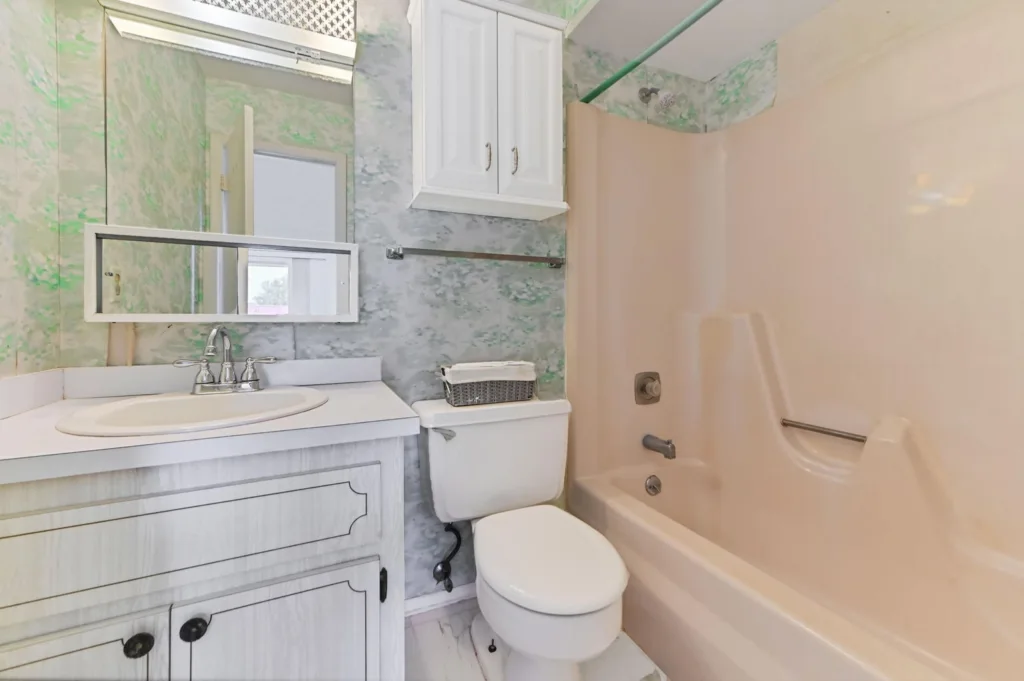 20260909143439479365000000-o - Lake Worth Homes - Lake Worth Homes White bathroom with a marble-green wall, a white vanity and sink, a rectangular mirror, and a toilet beside a beige bathtub with a grab bar and shower area.