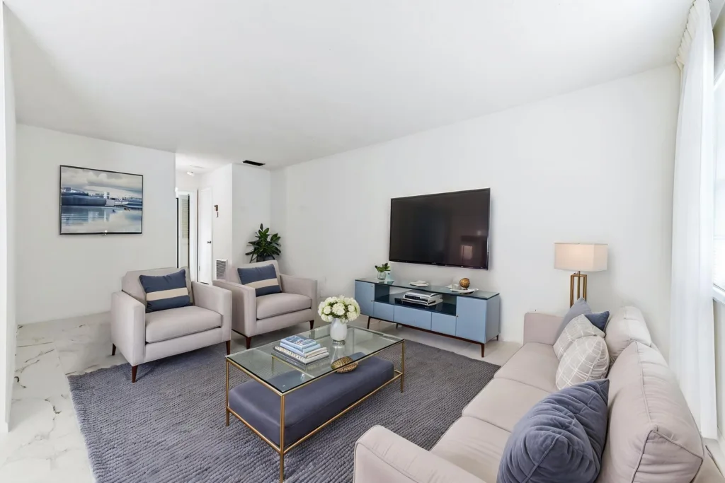 20260923173321872737000000-o 1 - Lake Worth Homes - Lake Worth Homes Bright living room with a wall-mounted TV, blue console, and pale sofas arranged around a glass coffee table on a gray rug.