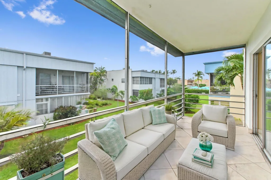 20260917220633270623000000-o - Lake Worth Homes - Lake Worth Homes Sunlit screened balcony with white wicker sofa and chair, teal pillows, and a glass-topped table overlooking a landscaped courtyard.