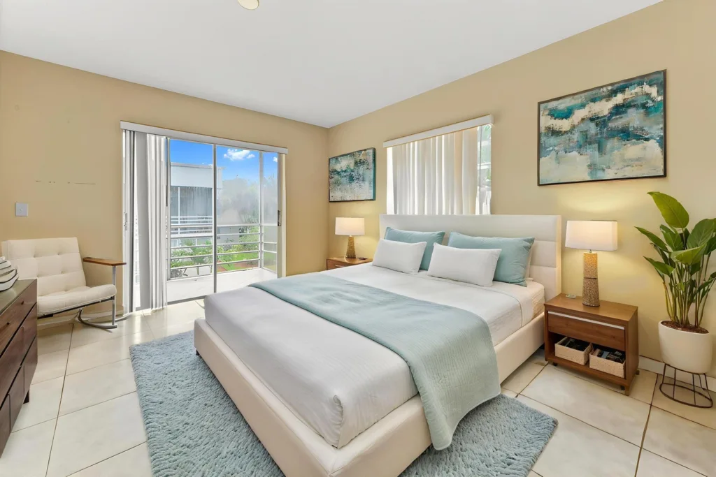 20260917220623762069000000-o - Lake Worth Homes - Lake Worth Homes Bright bedroom with a king bed, teal accents, two nightstands, lamps, and a sliding glass door to a balcony.