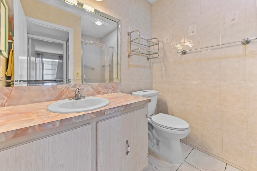 20260917212508924309000000-o - Lake Worth Homes - Lake Worth Homes Bathroom vanity with pink marble countertop, sink, and large wall mirror next to a toilet against beige tiled walls.