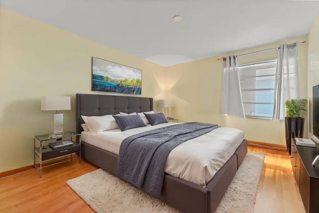 20260917220617612591000000-o - Lake Worth Homes - Lake Worth Homes Modern bedroom with a gray upholstered king bed, white bedding and blue-gray throw, flanked by glass nightstands and lamps.