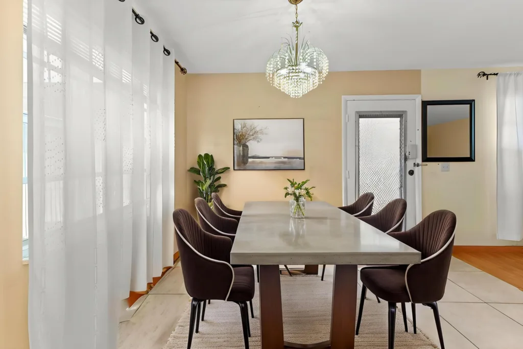 20260917220612015979000000-o - Lake Worth Homes - Lake Worth Homes Modern dining room: long concrete table with six brown upholstered chairs, chandelier, beige walls, and a vase with flowers on the table.
