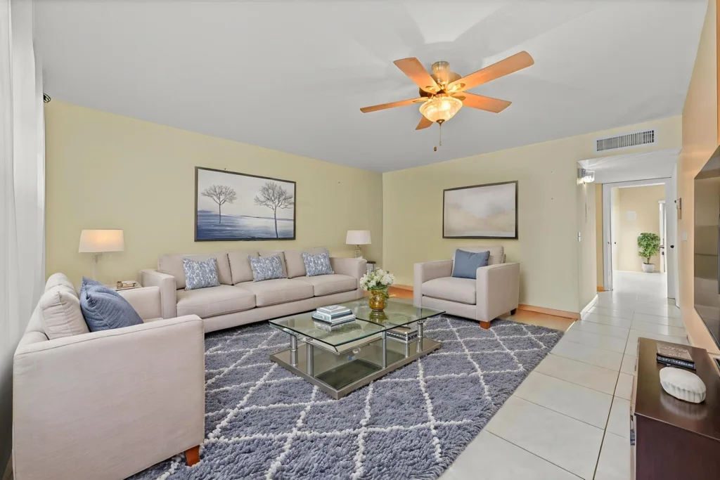 20260917220606472704000000-o - Lake Worth Homes - Lake Worth Homes Bright living room with beige sectional, matching armchairs, blue throw pillows, glass coffee table, and a plush gray rug with a white geometric pattern.