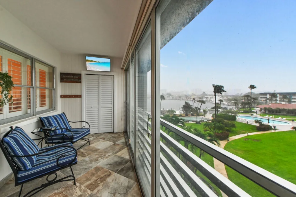 20260923233717735982000000-o - Lake Worth Homes - Lake Worth Homes Enclosed balcony with blue striped cushioned chairs and a small table, white shutters, and a view of a resort lawn, pool, and waterway.