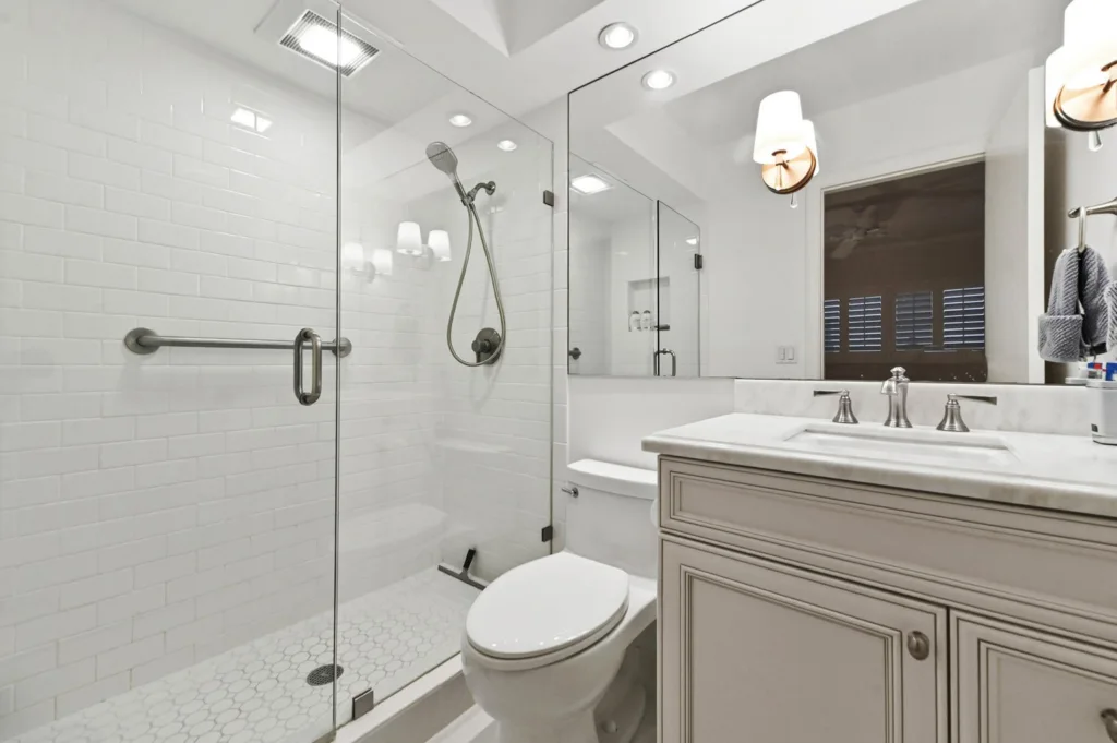 20260923233551743385000000-o - Lake Worth Homes - Lake Worth Homes Modern bathroom with a glass-enclosed shower, white subway-tile walls, a horizontal grab bar, and hexagonal floor tiles beside a white toilet and marble-top vanity with brushed-nickel fixtures.