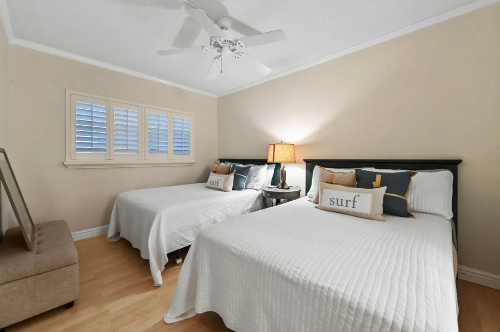 20260923233251784882000000-o - Lake Worth Homes - Lake Worth Homes Bedroom with two white-quirted beds, decorative pillows reading'surf', and a shared nightstand lamp between them on a beige wall. Window shutters on the left.
