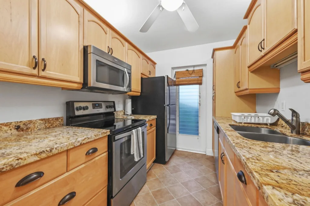 20260923233103440099000000-o - Lake Worth Homes - Lake Worth Homes Galley kitchen with light wood cabinets, granite countertops, stainless steel stove, microwave, and refrigerator. A sink and dish rack sit along the right counter.