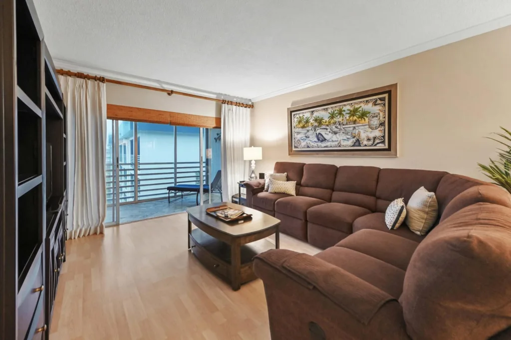20260923233150037837000000-o - Lake Worth Homes - Lake Worth Homes Living room with a brown sectional sofa, wooden coffee table, and palm-framed artwork above, beside a sliding balcony door.