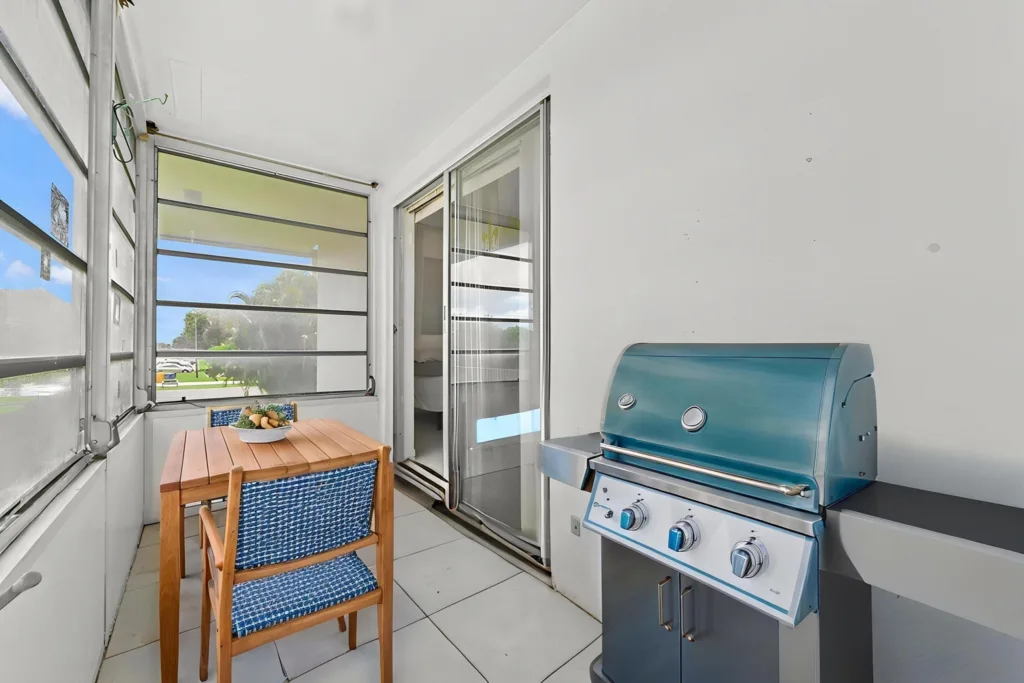 20260925152526360060000000-o - Lake Worth Homes - Lake Worth Homes Enclosed balcony with a wooden table and blue woven chairs beside a teal gas grill, sliding door to indoors.