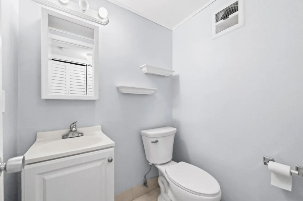 20260921183342059120000000-o - Lake Worth Homes - Lake Worth Homes Small bathroom with white sink, mirror cabinet, and toilet against light gray walls, plus a pair of shelves above the toilet