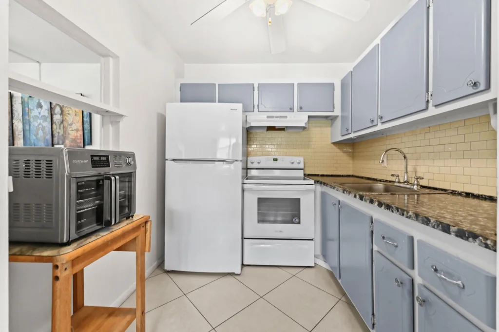 20260921183332898039000000-o - Lake Worth Homes - Lake Worth Homes Compact kitchen with light-blue cabinets, beige tile backsplash, white refrigerator and stove, granite countertop.