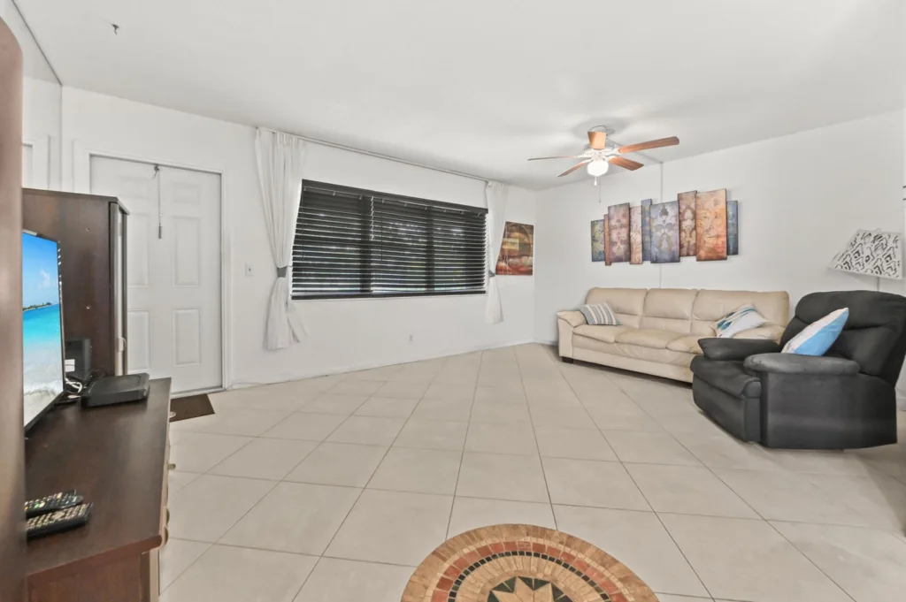 20260925150741792617000000-o - Lake Worth Homes - Lake Worth Homes Bright living room with a beige sofa and black recliner, ceiling fan, wall art, and a large window with blinds; TV on a wooden cabinet to the left.