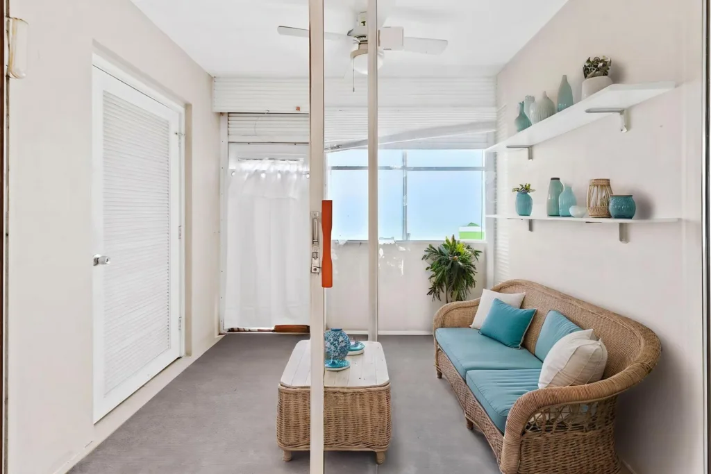 20260924144223817831000000-o - Lake Worth Homes - Lake Worth Homes Bright sunroom with a wicker loveseat featuring teal cushions, white shelves with turquoise vases, and a potted plant near glass doors.