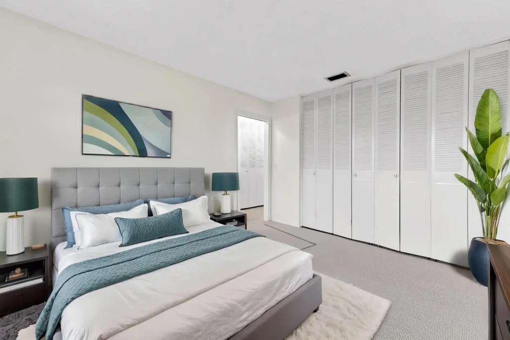 20260924144120928991000000-o - Lake Worth Homes - Lake Worth Homes Modern bedroom with gray upholstered king bed, teal accents, two nightstands and teal lamps, abstract wall art, white walls, and a row of white closet doors.