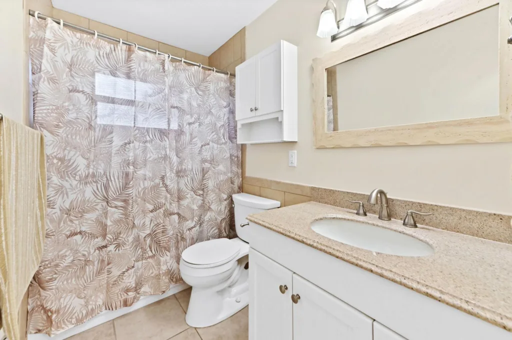 20260924144051292029000000-o - Lake Worth Homes - Lake Worth Homes Bathroom with a light granite countertop, white cabinets, and a framed mirror; palm-leaf shower curtain panels the tub/shower area, with a white toilet beside the vanity.