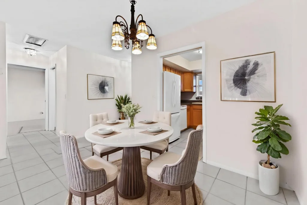 20260924144007871857000000-o - Lake Worth Homes - Lake Worth Homes Round white dining table with four upholstered chairs in a bright dining area; chandelier overhead, kitchen with wooden cabinets and white fridge visible in the background; a large plant and framed abstract art decorate the walls.