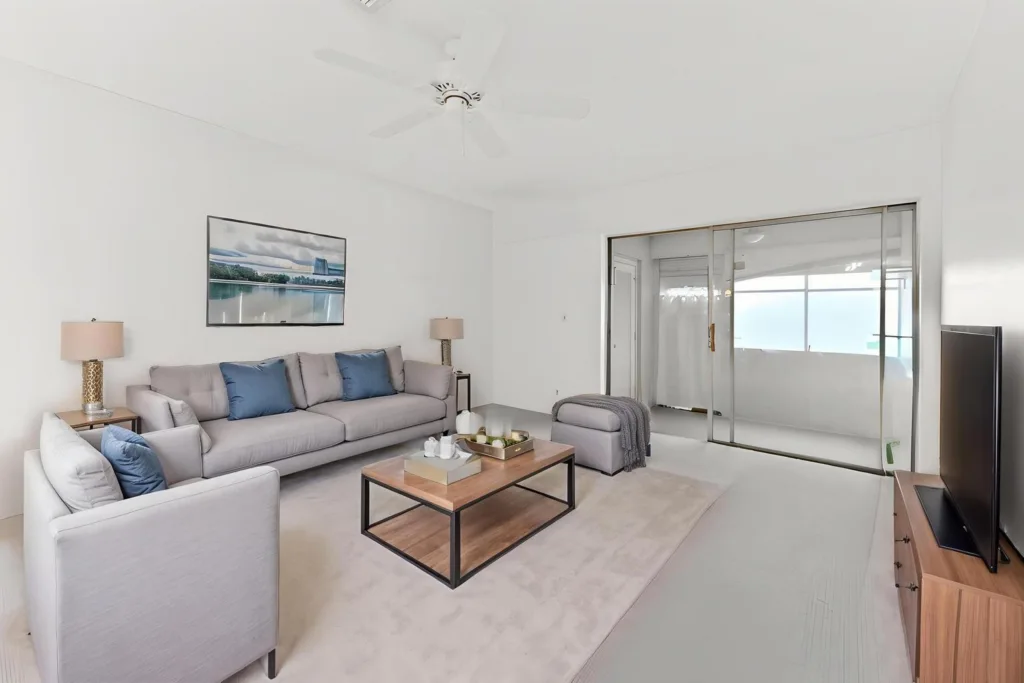 20260924143943104738000000-o - Lake Worth Homes - Lake Worth Homes Bright, minimalist living room with a light gray sofa and armchairs, blue cushions, a wooden coffee table, and a wall-mounted art piece above the couch far wall source light from windows off-screen.