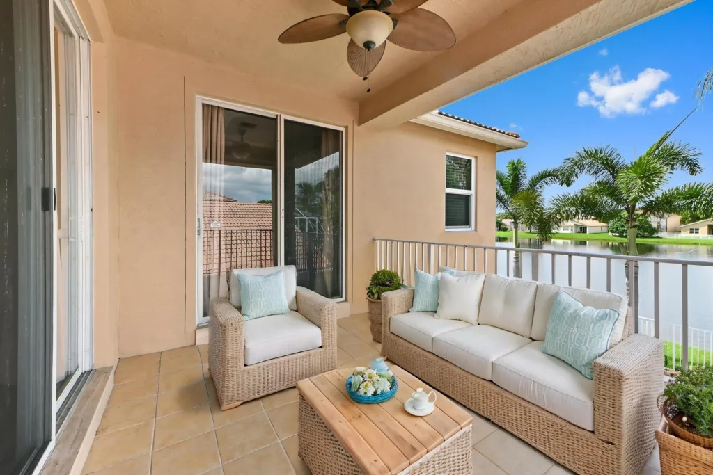 20260922221724163104000000-o - Lake Worth Homes - Lake Worth Homes Outdoor balcony seating area with beige wicker sofa and chair, cream cushions, and turquoise pillows overlooking a lake.