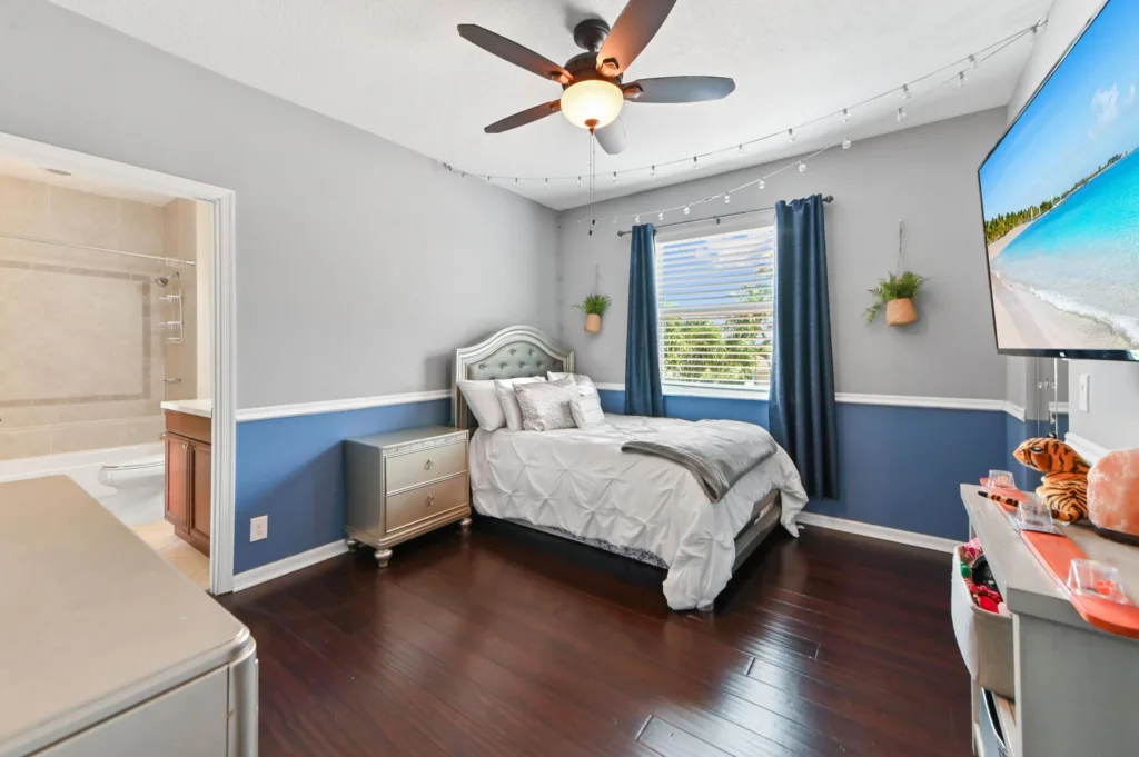 20260922220408187112000000-o - Lake Worth Homes - Lake Worth Homes Bedroom with gray and blue walls, queen bed with tufted headboard, string lights, and a ceiling fan; window with blue curtains, dresser nearby, and a TV showing a beach scene. There’s an en-suite bathroom visible to the left.