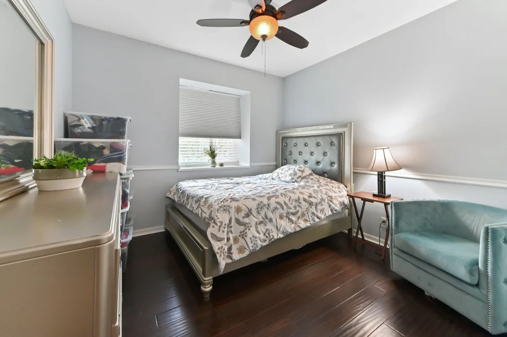 20260922220435870505000000-o - Lake Worth Homes - Lake Worth Homes Bedroom with a gray tufted headboard bed and floral bedding, teal chair, bedside lamp, and clear plastic storage drawers along the left wall.