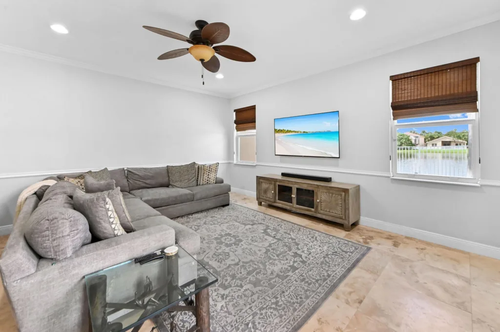 20260922220157900120000000-o - Lake Worth Homes - Lake Worth Homes Light gray living room with an L-shaped sectional, patterned cushions, a wall-mounted TV above a wooden console, and two windows with brown blinds.