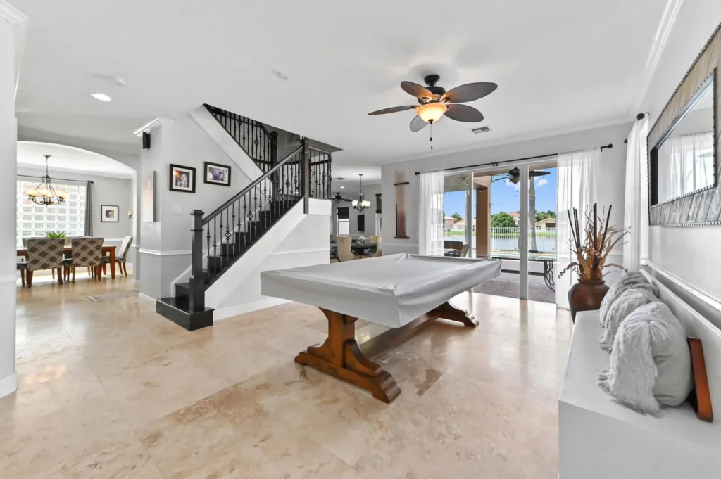 20260922220110089779000000-o 1 - Lake Worth Homes - Lake Worth Homes Open-concept living room with a covered pool table, black iron staircase railing, and sliding doors to a sunny patio overlooking a lake garden area.