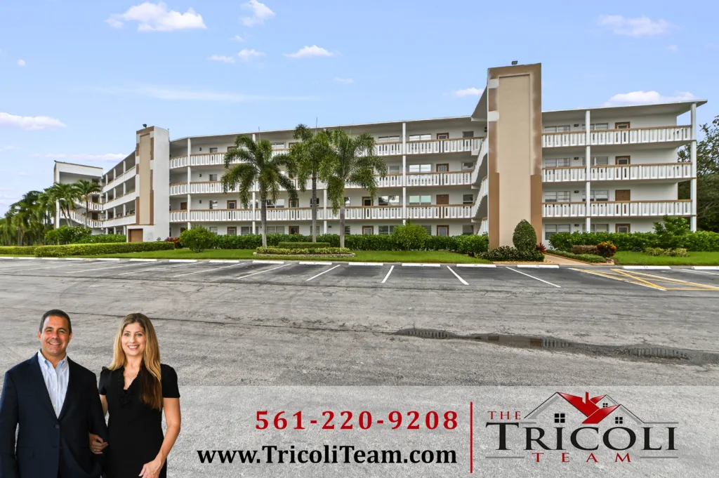 Homes for Sale in Cornwall at Century Village Boca Raton FL 33434 - Lake Worth Homes - Lake Worth Homes Two casually dressed professionals stand at the edge of a large parking lot in front of a multi-story apartment building with palm trees and white railings under a blue sky.