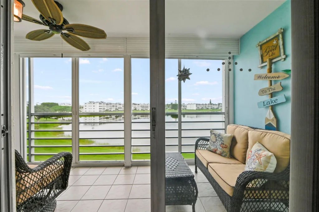 20260929155558643505000000-o - Lake Worth Homes - Lake Worth Homes Balcony living area with wicker sofa and chairs, turquoise wall sign'Welcome To The Lake', overlooking a lake and buildings beyond.