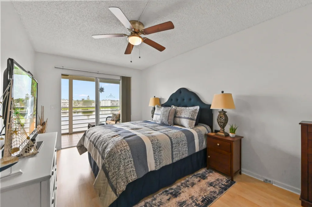 20260929155426845353000000-o - Lake Worth Homes - Lake Worth Homes Bright bedroom with a queen bed, blue tufted headboard, matching bedding, and balcony doors opening to a river view.
