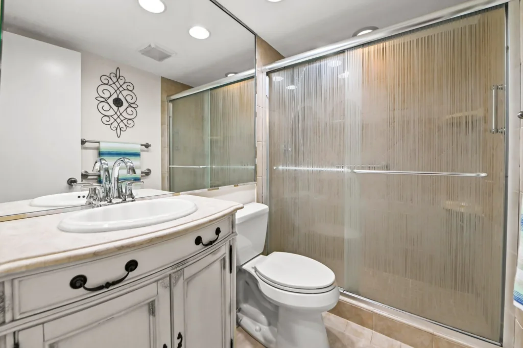 20260929155328440368000000-o - Lake Worth Homes - Lake Worth Homes Modern bathroom with white vanity, marble countertop, chrome faucet, and toilet beside a frosted-glass shower enclosure.