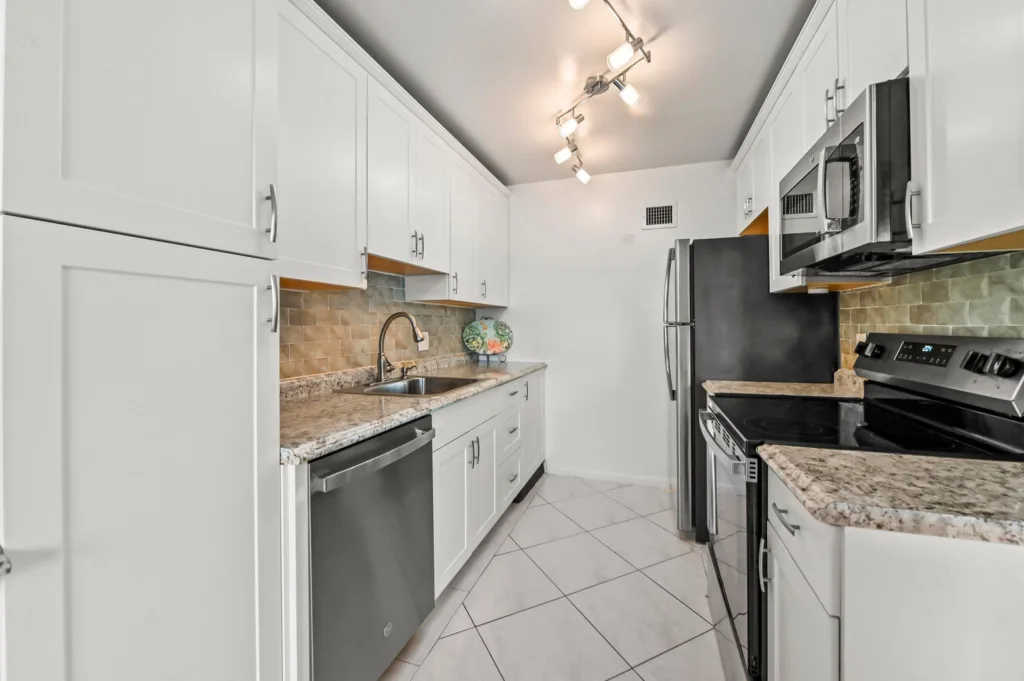 20260929155305415329000000-o - Lake Worth Homes - Lake Worth Homes Bright, modern galley kitchen with white cabinets, granite countertops, beige tile backsplash, and stainless steel appliances (refrigerator, microwave, oven, and dishwasher). Track lighting overhead.