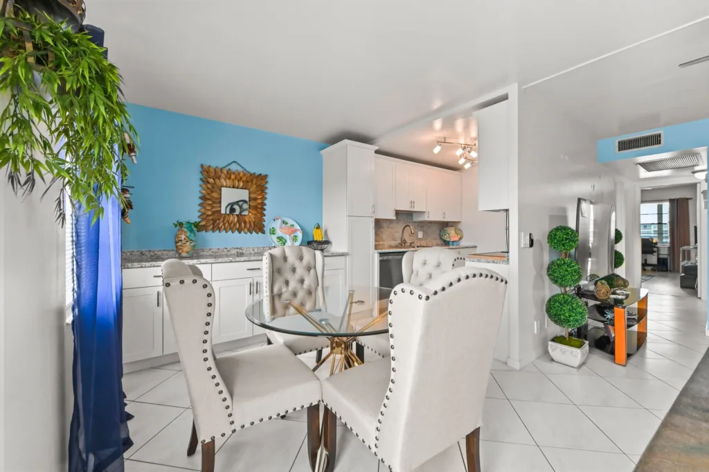 20260929155232781476000000-o - Lake Worth Homes - Lake Worth Homes Open-concept kitchen and dining room with a blue accent wall, white cabinets, and a glass round dining table surrounded by tufted cream chairs; decorative sunburst mirror and potted plants add color.