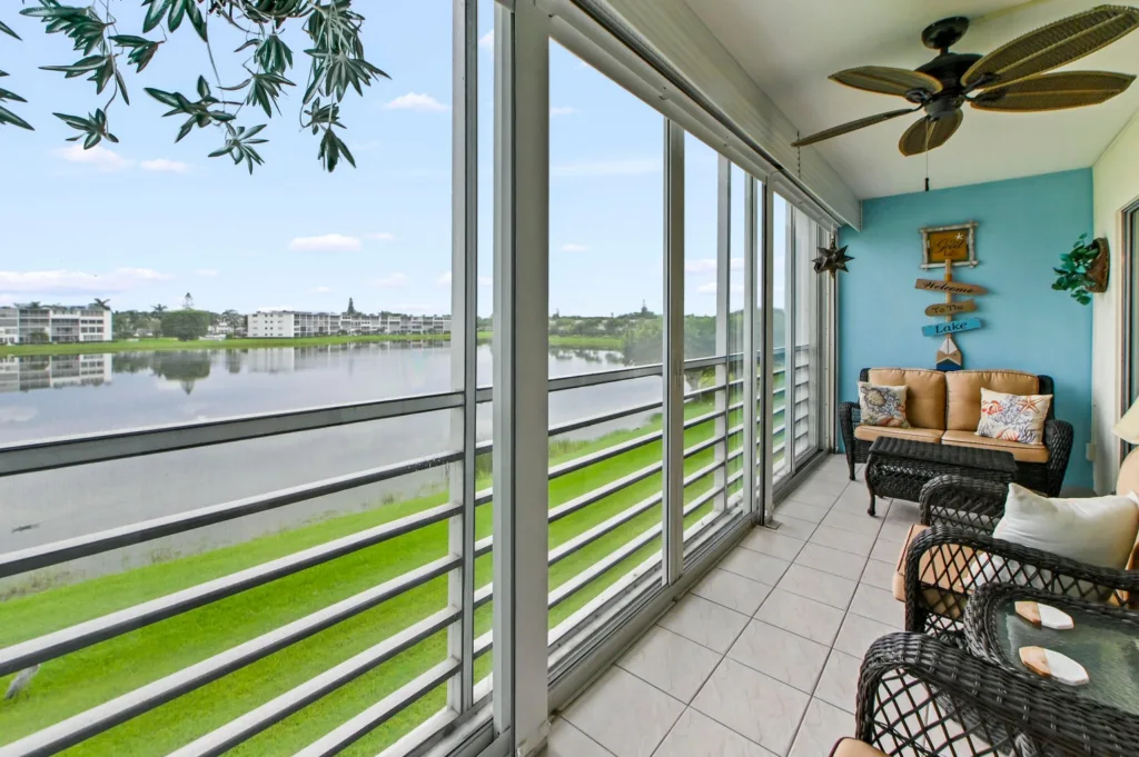 20260929155543095386000000-o - Lake Worth Homes - Lake Worth Homes Enclosed balcony with wicker loveseat and chair, beige cushions, turquoise wall, and a view of the lake across the water.
