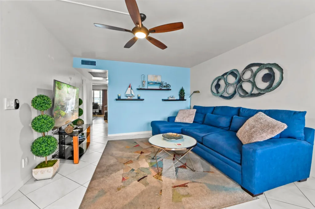 20260929155104077352000000-o - Lake Worth Homes - Lake Worth Homes Cozy living room with a bright blue sectional sofa, round glass coffee table, and a multicolored rug underfoot, plus circular metal wall art above the sofa.