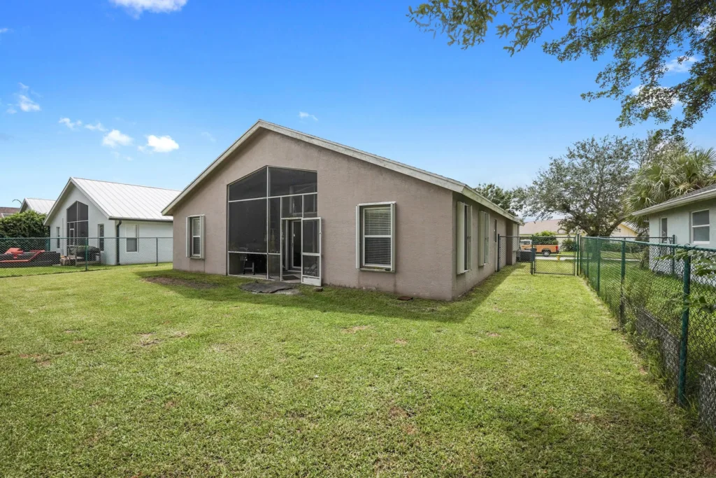 20260924162313285257000000-o - Lake Worth Homes - Lake Worth Homes Backyard of a single-story beige house with a screened-in patio, green lawn, and chain-link fencing; neighboring white houses nearby under a blue sky.